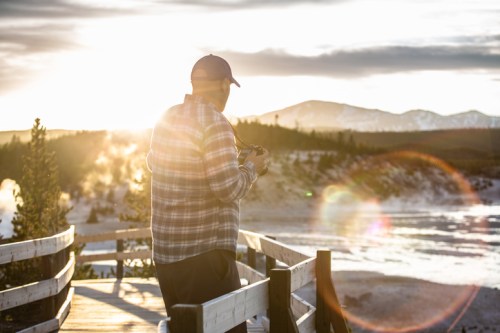 A man stands on a wooden walkway, holding a camera, with a sunset and mountains in the background.