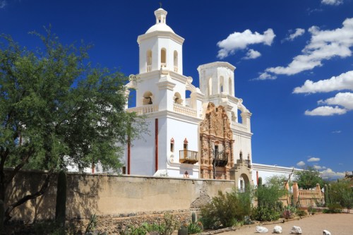 Historic white building with twin bell towers, ornate facade, and lush greenery under a bright blue sky.