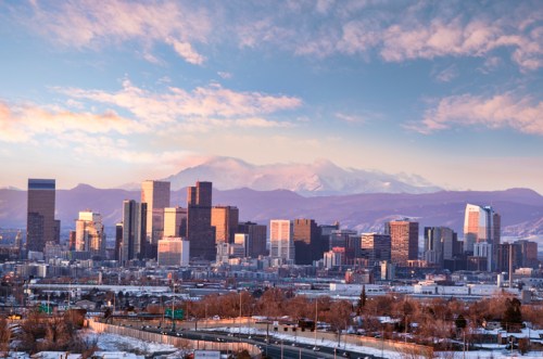 A panoramic view of a city skyline with mountains in the background, under a partly cloudy sky.