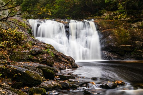 A serene waterfall cascades over rocks, surrounded by autumn foliage and a calm pool below.
