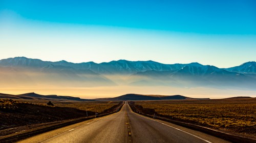 A long, empty road stretches towards distant mountains under a clear blue sky, with soft light illuminating the landscape.