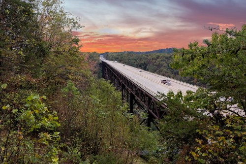 A highway bridge spans a forested valley at sunset, with cars traveling on the road beneath a colorful sky.