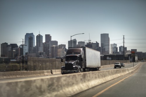 A truck drives on a highway with a city skyline in the background under a clear sky.