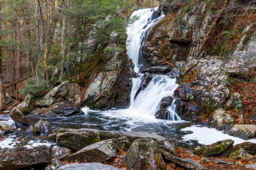 A serene waterfall cascades over rocky terrain, surrounded by lush greenery and autumn leaves.