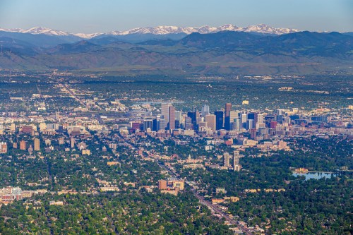 Aerial view of Denver's skyline with mountains in the background, surrounded by greenery and urban development.