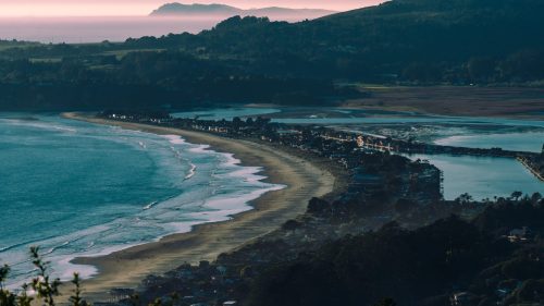 A scenic coastal view featuring a sandy beach, calm waters, and distant hills under a soft sunset glow.