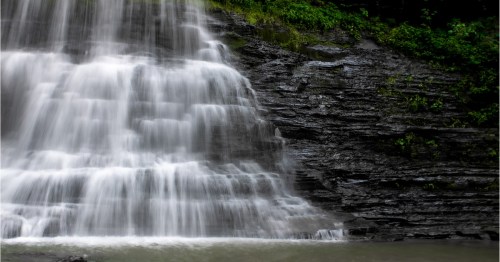 A cascading waterfall flows over dark, rocky cliffs surrounded by lush greenery.
