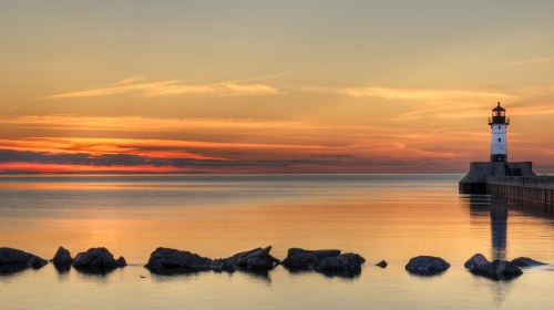 A serene sunset over calm waters, featuring a lighthouse on a pier and rocky shoreline.