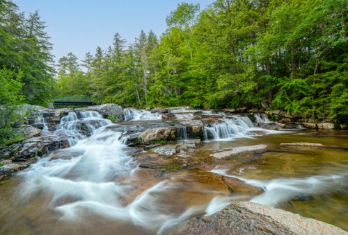A serene waterfall cascades over rocks, surrounded by lush green trees and a clear blue sky.
