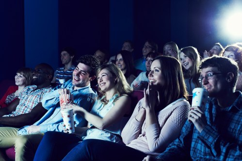 A diverse group of people enjoying a movie in a theater, laughing and holding drinks.