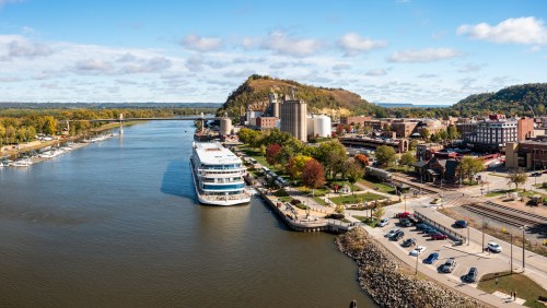 A scenic view of a river with a boat docked, surrounded by autumn trees and buildings under a blue sky.