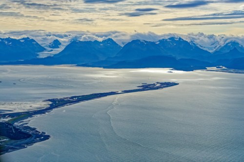 Aerial view of a coastal landscape with mountains, water, and a peninsula under a cloudy sky.