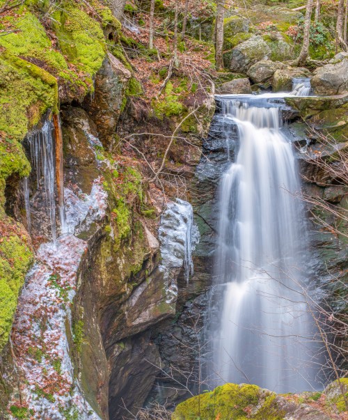 A serene waterfall cascades over moss-covered rocks, surrounded by trees and autumn leaves.