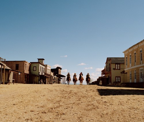 Four cowboys on horseback ride down a dusty street lined with old western-style buildings under a clear blue sky.