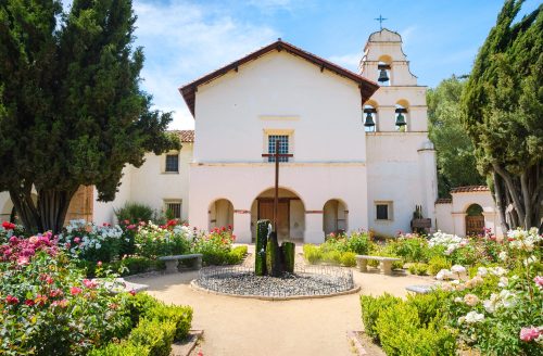 A historic building surrounded by a vibrant garden filled with roses and greenery, featuring a bell tower in the background.