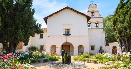 A historic building with a bell tower, surrounded by colorful flowers and greenery, under a clear blue sky.