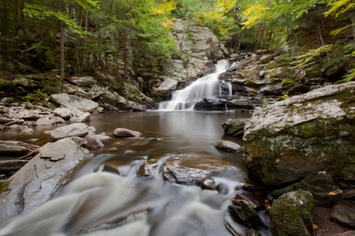 A serene waterfall cascades over rocks into a calm pool, surrounded by lush green trees and autumn foliage.