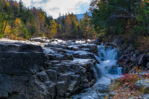 A serene waterfall cascades over rocky terrain, surrounded by autumn foliage and trees under a cloudy sky.