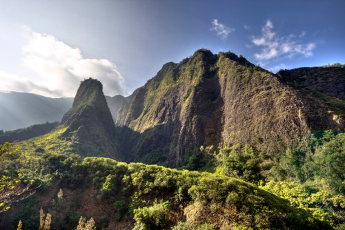 Lush green mountains rise under a clear blue sky, with sunlight illuminating the rocky peaks and valleys.