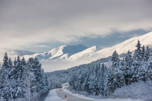 A snowy road winds through a forest, with majestic mountains in the background under a cloudy sky.
