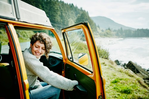 A person with curly hair smiles while sitting in the open door of a yellow vehicle by a scenic coastal landscape.