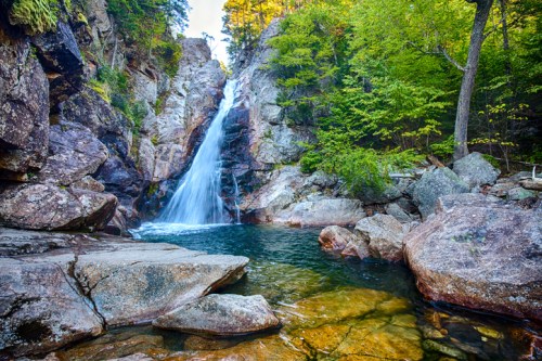 A serene waterfall cascades over rocky cliffs into a clear pool, surrounded by lush green trees and vibrant foliage.