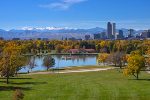 A scenic view of a park with a lake, surrounded by autumn trees and a city skyline against a backdrop of snow-capped mountains.