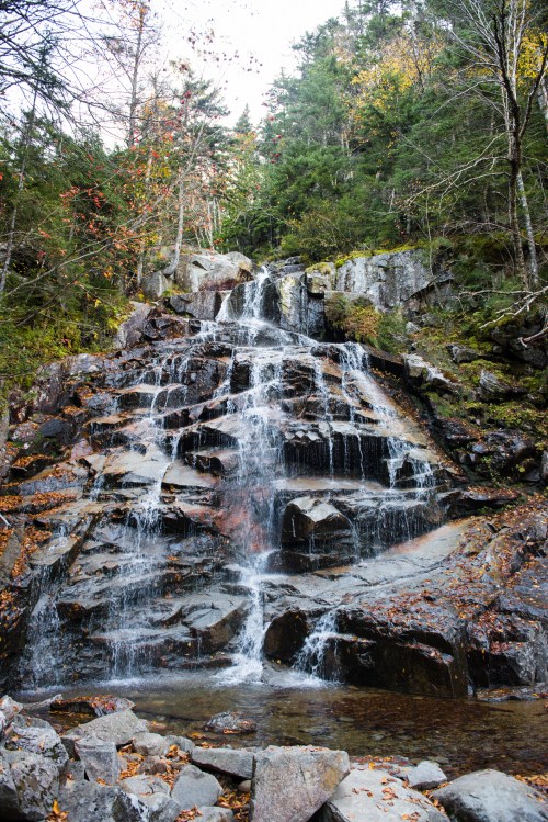 A serene waterfall cascading over rocky terrain, surrounded by autumn foliage and lush green trees.
