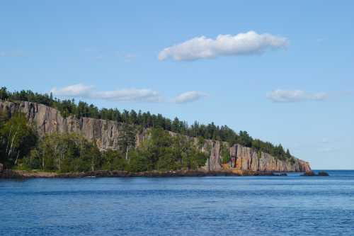 A rocky shoreline with lush greenery under a clear blue sky and fluffy clouds, reflecting in calm waters.