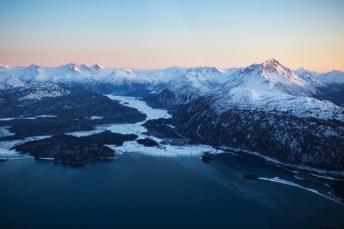 Aerial view of snow-capped mountains and a frozen lake at sunset, with soft pastel colors in the sky.