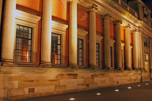 Illuminated building facade at night, featuring tall columns and large windows along a stone wall.