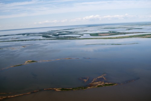 Aerial view of a vast, tranquil water landscape with islands and lush greenery under a blue sky with scattered clouds.