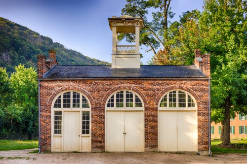 Historic brick building with arched windows and a bell tower, surrounded by trees and mountains in the background.
