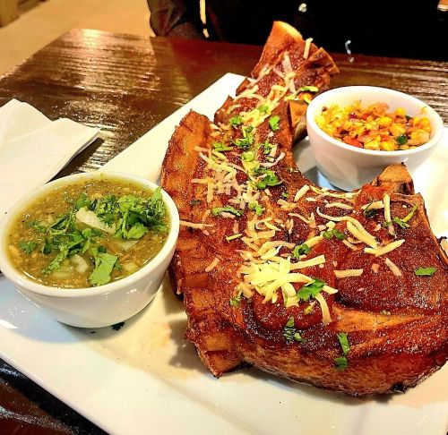 A plate featuring a large grilled meat chop topped with cheese, accompanied by green sauce and a side of corn salad.