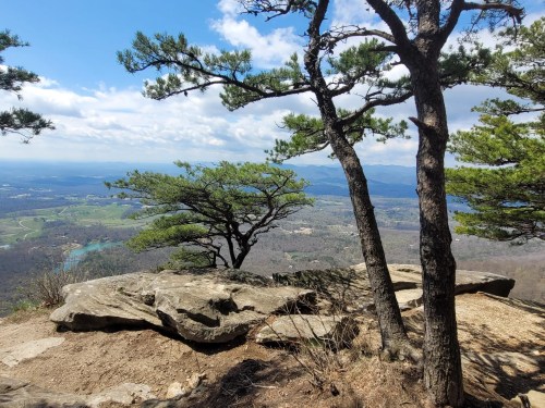 A scenic view from a rocky overlook, featuring trees and rolling hills under a partly cloudy sky.