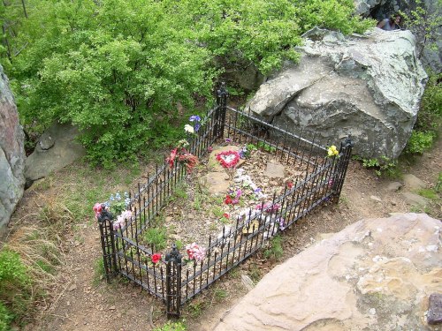 A small, fenced grave adorned with flowers, surrounded by rocks and greenery in a natural setting.