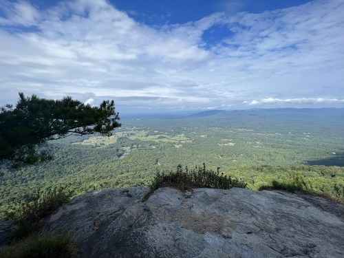 A panoramic view of lush green valleys and distant mountains under a partly cloudy sky from a rocky overlook.