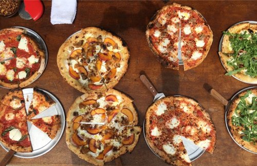 A variety of pizzas on a wooden table, including classic, fruit-topped, and arugula-topped options.