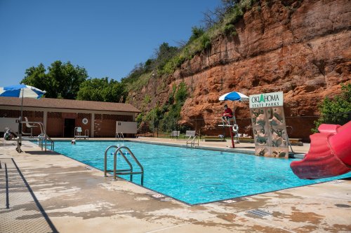 A sunny pool area with a lifeguard, water slide, and rock formations in the background at Oklahoma State Parks.