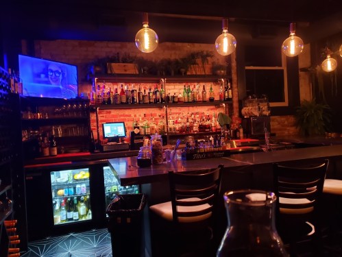A dimly lit bar with shelves of bottles, a TV screen, and warm pendant lights illuminating the space.