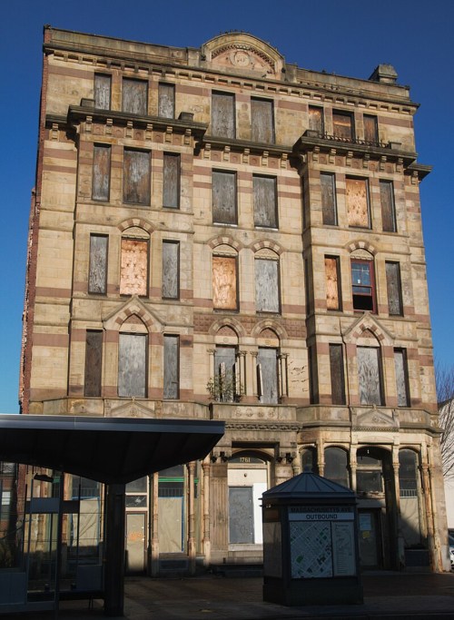 An abandoned, multi-story building with boarded windows and ornate architectural details against a clear blue sky.