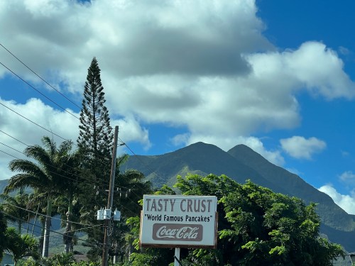 Sign for "Tasty Crust" featuring "World Famous Pancakes," with mountains and palm trees in the background.