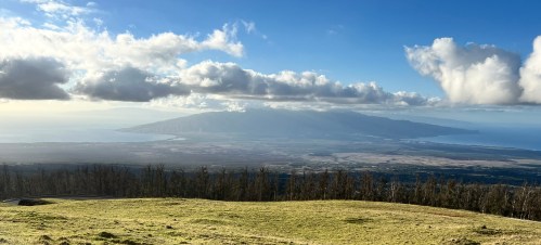 A panoramic view of a lush landscape with rolling hills and a distant island under a partly cloudy sky.