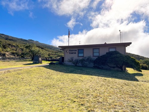 A visitor center building surrounded by grass and trees, with a flagpole and cloudy sky in the background.