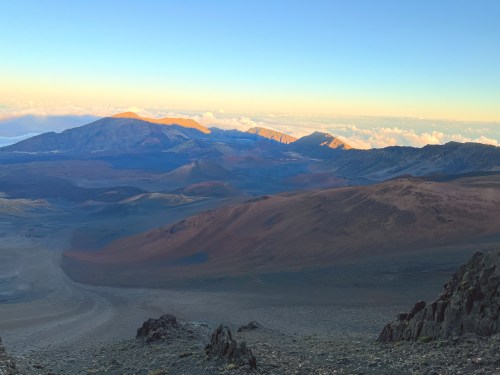A panoramic view of rugged mountains and valleys under a colorful sky at sunrise, with clouds in the distance.