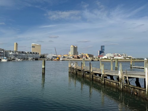 A calm waterfront scene with a wooden pier, city skyline, and blue sky with scattered clouds in the background.