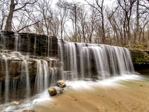 A serene waterfall cascades over rocks into a sandy area, surrounded by bare trees in a tranquil forest setting.