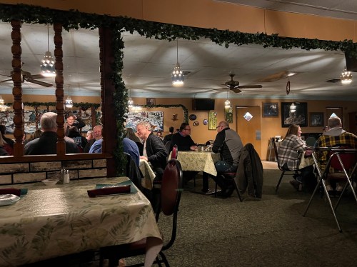 A cozy restaurant interior with diners at tables, warm lighting, and rustic decor featuring plants and wooden accents.