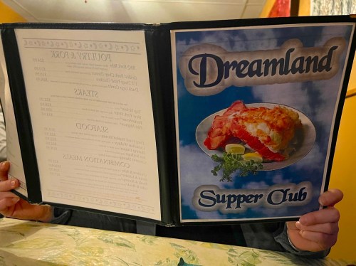 A person holds a menu for "Dreamland Supper Club," featuring a dish of lasagna on the cover.