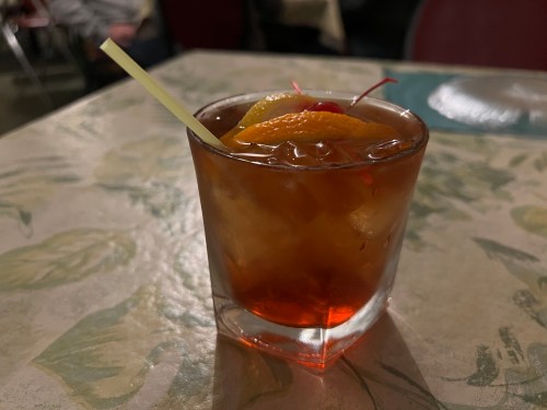 A close-up of a cocktail in a glass, garnished with orange slices and a cherry, resting on a patterned table.
